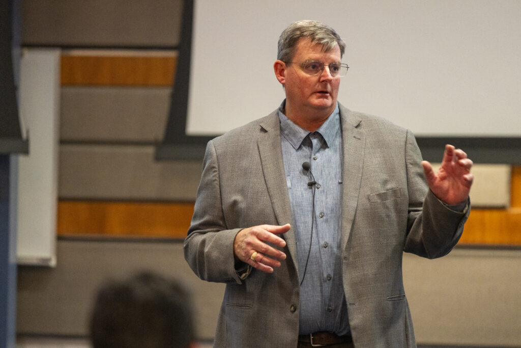 A man in a grey suit and blue shirt confidently presenting to an audience.