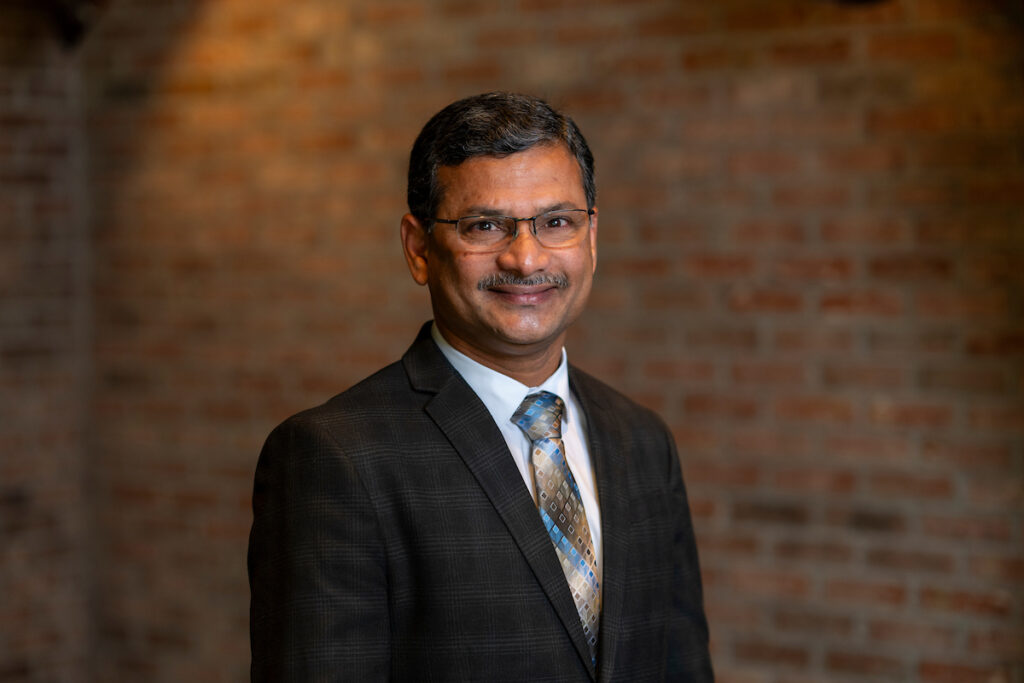 A man, Srinivasulu Ale, Ph.D., in a jacket and tie in front of a brick wall background