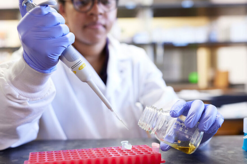 Student wearing protective lab gear and filling a test tube using a pipette.