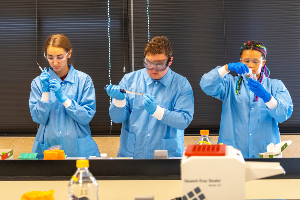 Three students wearing lab coats and safety glasses conducting an experiment in a lab.
